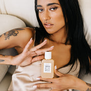 Model with black hair, white nail polish, facial piercings, and dragon tattoo on arm wears beige satin tank top while laying down on off-white couch and holding Vanilla Milk 50ml Eau De Parfum with both hands.