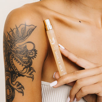 Model in strapless beige top with dragon tattoo and white nail polish holding Vanilla Milk 10ml Eau De Parfum Travel Spray below collarbone on white background.