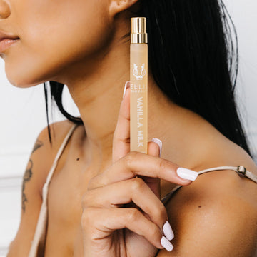 Model with black hair, white nail polish, and tattoo on arm wears a beige satin tank top and holds Vanilla Milk 10ml Eau De Parfum Travel Spray near shoulder, against white background.