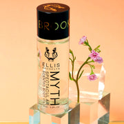 Myth Spectacular Scented Body Oil 95ml on glass block in front of pink flowers, glass block, and glass cylinder, on orange background.