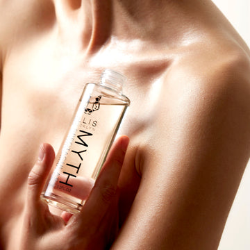 Model holding Myth 95ml Spectacular Scented Body Oil with out cap, near collarbone, on white background.