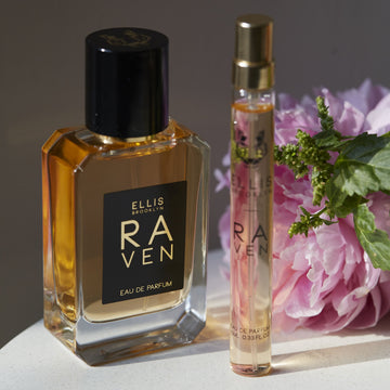 Raven 50ml Eau De Parfum on white table next to Raven 10ml Eau De Parfum Travel spray, pink flower, and green leaves, on brown background.