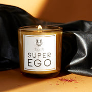 Superego 6.5 oz. Terrific Scented Candle with wick lit on orange backdrop with saffron and black leather.