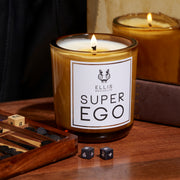 Superego 6.5 oz. Terrific Scented Candle on wooden table next to game with dice and dice on table, in front of brown book, mirror, and black fabric.