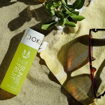 Sun Fruit Golden Glistening Scented Body Oil 95ml on sand near yellow and white striped towel, green plant, and brown sunglasses.
