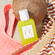 Sun Fruit 50ml Eau De Parfum resting on coconut and pink bathing suit top in sand next to wicker strap and orange and white striped shirt.