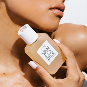 Model with white nail polish and diamond stud above lip holding Vanilla Milk 50ml Eau De Parfum near jaw, on white background.